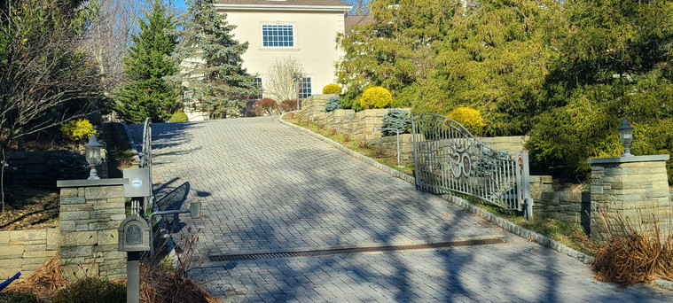 Paver driveway with iron gate, stone retaining walls, and layered evergreen plantings